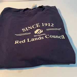 K&ouml;nnte beinhalten: Ein marineblaues T-Shirt mit wei&szlig;em Siebdrucktext, der SINCE 1912 lautet, einem Girl Scouts Logo und den Worten Red Lands Council. Das T-Shirt hat einen Rundhalsausschnitt und ein sichtbares wei&szlig;es Stoffetikett am Kragenbereich.