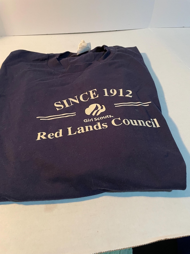 K&ouml;nnte beinhalten: Ein marineblaues T-Shirt mit wei&szlig;em Siebdrucktext, der SINCE 1912 lautet, einem Girl Scouts Logo und den Worten Red Lands Council. Das T-Shirt hat einen Rundhalsausschnitt und ein sichtbares wei&szlig;es Stoffetikett am Kragenbereich.
