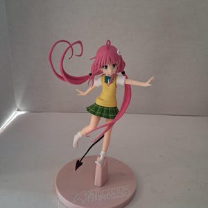 May include: A figurine of a female anime character with pink hair and green eyes. She wears a yellow vest, white shirt, and green plaid skirt. The figure is on a pink base with text and a devil tail.