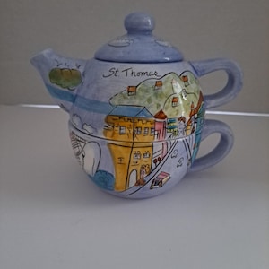 May include: A blue and yellow ceramic teapot with a painted scene of St. Thomas. The teapot has a lid and a handle. The scene includes buildings, a road, and clouds.