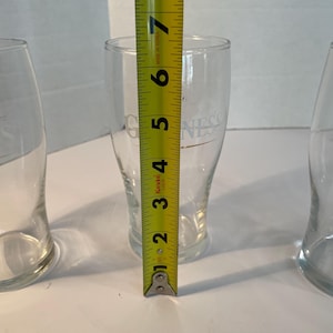 May include: Three clear pint glasses with the word "Guinness" printed on them. The center glass is measured with a yellow tape measure, showing a height of approximately 6 inches.