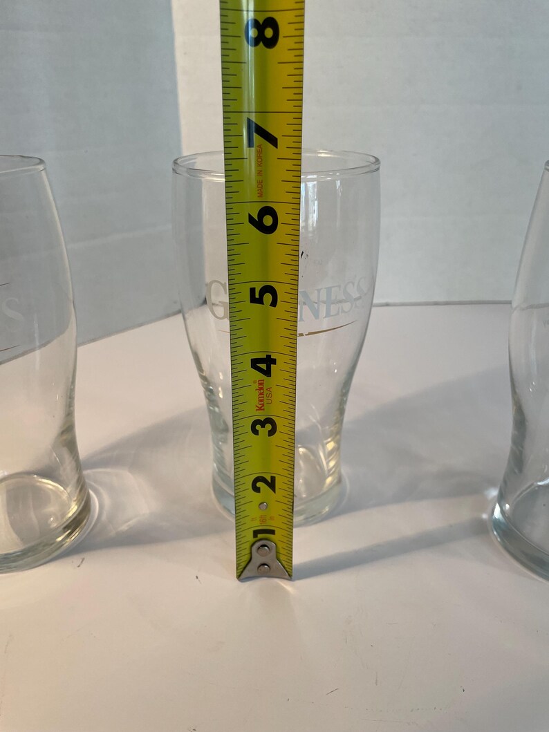 May include: Three clear pint glasses with the word "Guinness" printed on them. The center glass is measured with a yellow tape measure, showing a height of approximately 6 inches.