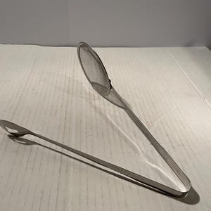 May include: A silver-colored metal kitchen tong featuring a circular mesh strainer on one end and a solid, slightly curved metal paddle on the other. The two arms are connected at a hinge, allowing the tool to open and close for gripping food.