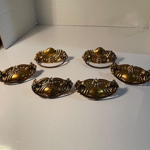 May include: Six antique-style drawer pulls with a gold finish. Each pull has an ornate, curved design with a decorative handle. The pulls are arranged on a white surface.