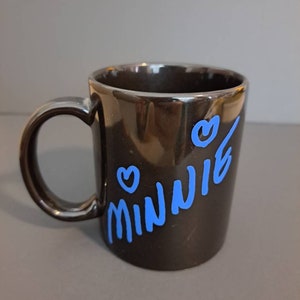 Peut inclure: Mug en c&eacute;ramique noire avec le nom "Minnie" en bleu, accompagn&eacute; de deux coeurs bleus. La tasse a une finition brillante et une anse incurv&eacute;e.