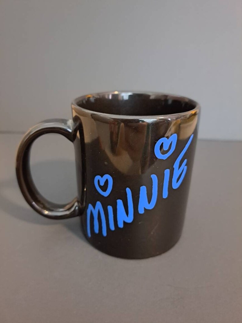 Peut inclure: Mug en c&eacute;ramique noire avec le nom "Minnie" en bleu, accompagn&eacute; de deux coeurs bleus. La tasse a une finition brillante et une anse incurv&eacute;e.