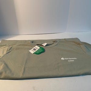 May include: A sage green short-sleeved t-shirt featuring the white text Girl Scouts Leader on the front right chest. A green clover-shaped tag is attached to the collar, and a white rectangular tag is tucked inside the neckline of the shirt.