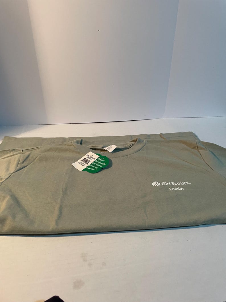 May include: A sage green short-sleeved t-shirt featuring the white text Girl Scouts Leader on the front right chest. A green clover-shaped tag is attached to the collar, and a white rectangular tag is tucked inside the neckline of the shirt.