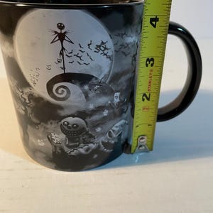 May include: A black ceramic mug featuring a grayscale illustration of characters from The Nightmare Before Christmas, including Jack Skellington and Oogie Boogie. A yellow Stanley measuring tape is placed against the side, showing a height of 5 inches.