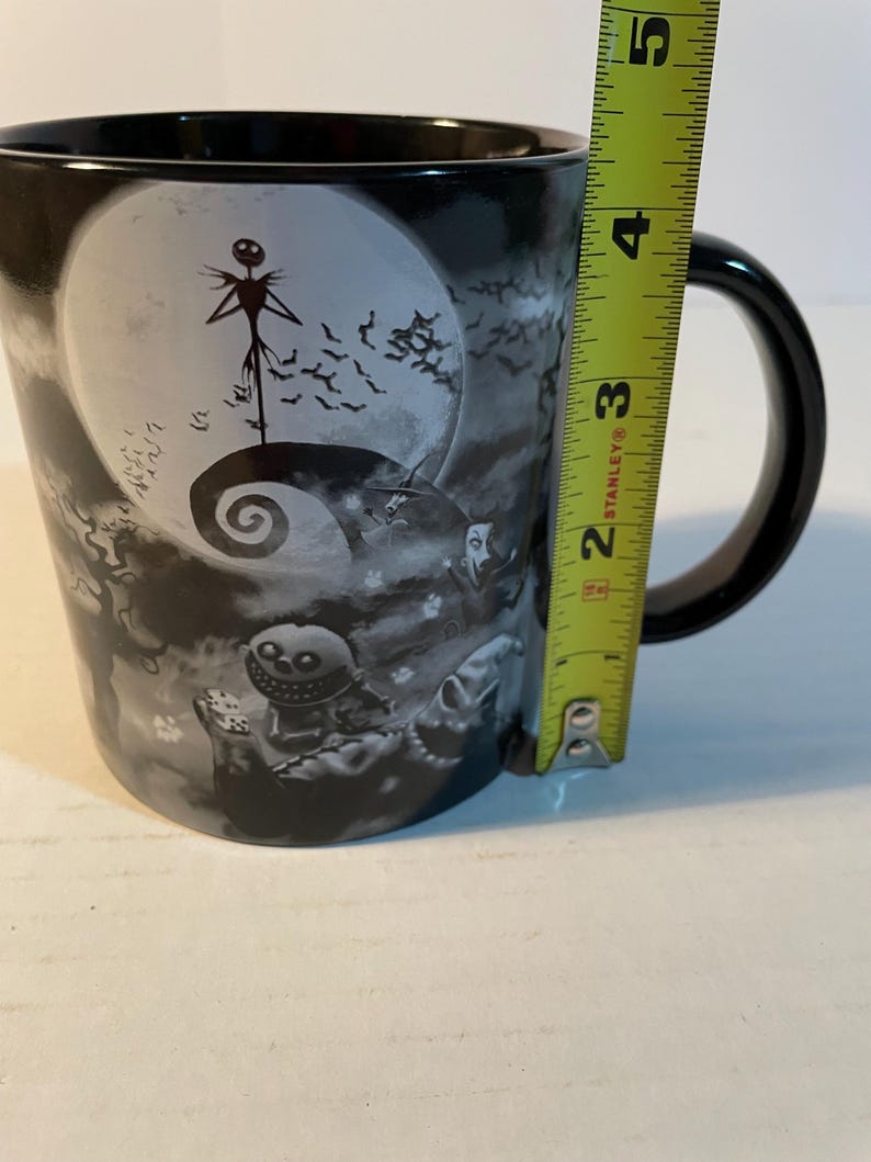 May include: A black ceramic mug featuring a grayscale illustration of characters from The Nightmare Before Christmas, including Jack Skellington and Oogie Boogie. A yellow Stanley measuring tape is placed against the side, showing a height of 5 inches.