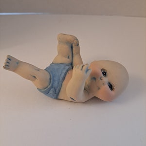 May include: A ceramic figurine of a baby wearing a blue diaper, lying on its back with its legs raised in the air. The baby has painted eyes and rosy cheeks.