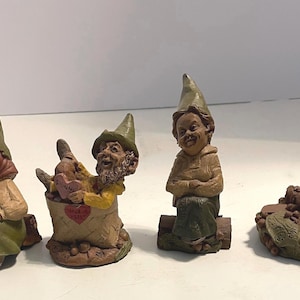 May include: Four small gnome figurines with pointed hats. The first wears a green hat and skirt. The second sits in a basket holding a pink heart. The third sits on a log wearing a green hat. The fourth sits on a base with a turtle and a red hat.