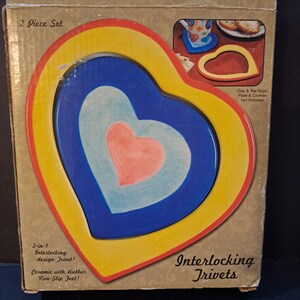 May include: A set of two interlocking heart-shaped trivets in yellow, red, blue, and light blue. The trivets are made of ceramic with rubber non-slip feet. The packaging says "Interlocking Trivets - 2 Piece Set".