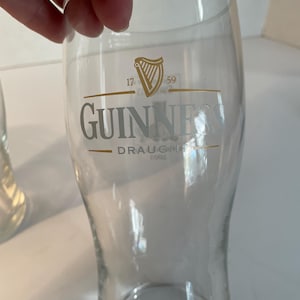May include: A clear, tulip-shaped Guinness pint glass. The glass features the Guinness logo in silver, with a gold harp emblem and the text "17 59". The words "Guinness Draught" are also visible.