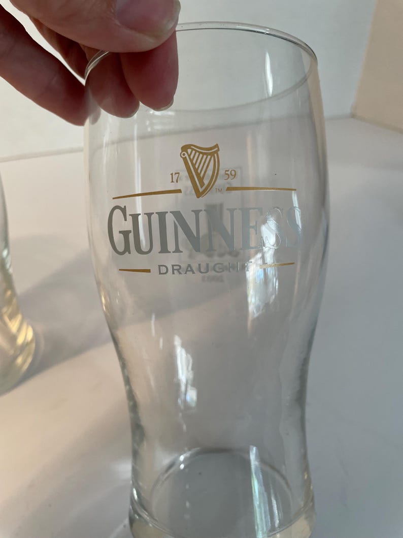 May include: A clear, tulip-shaped Guinness pint glass. The glass features the Guinness logo in silver, with a gold harp emblem and the text "17 59". The words "Guinness Draught" are also visible.