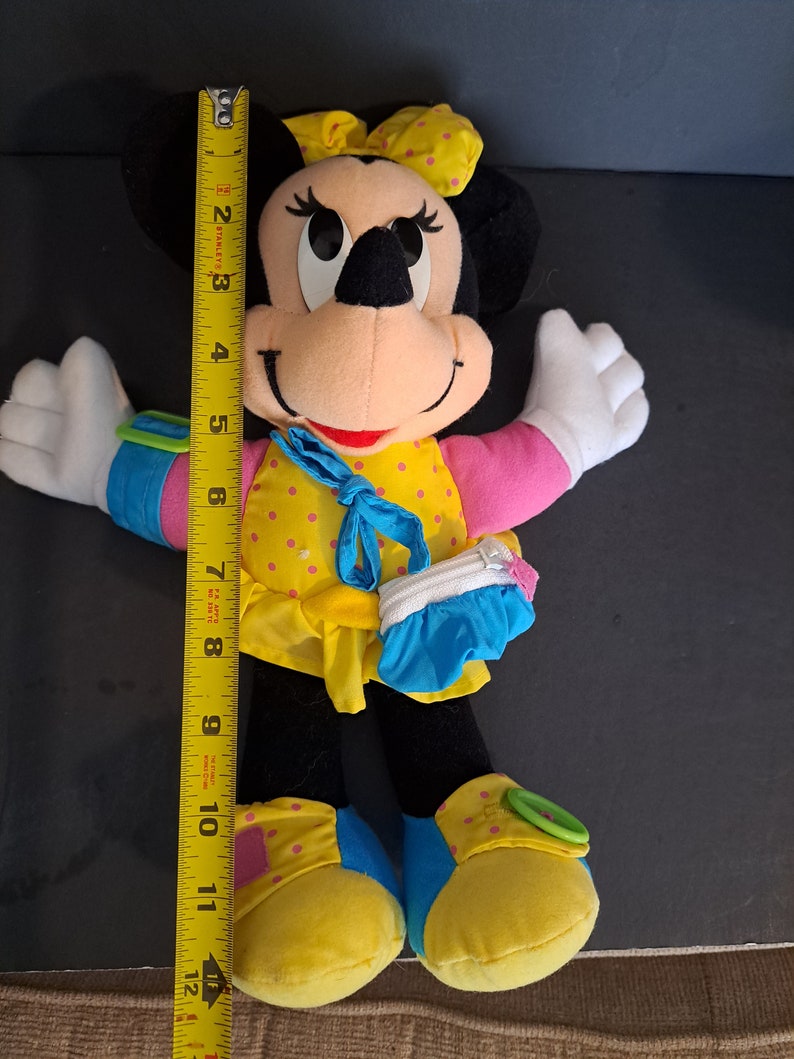 May include: A plush Minnie Mouse toy wearing a yellow dress with pink polka dots, a blue purse, and yellow shoes with pink polka dots. The toy has black ears and a red bow.