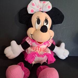 May include: A pink and white polka dot bow sits atop the head of a plush Minnie Mouse toy. She is wearing a pink and white striped dress with a pink tulle skirt. The toy is sitting with its legs crossed and has pink shoes.