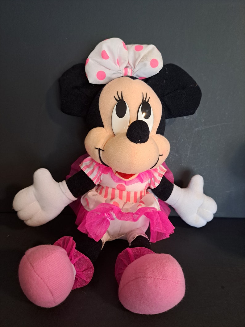 May include: A pink and white polka dot bow sits atop the head of a plush Minnie Mouse toy. She is wearing a pink and white striped dress with a pink tulle skirt. The toy is sitting with its legs crossed and has pink shoes.