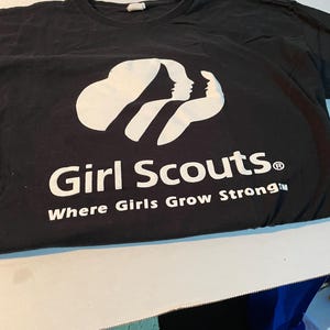 May include: A black short-sleeved t-shirt featuring a white graphic of three stylised female silhouettes. Below the graphic, the text reads Girl Scouts and Where Girls Grow Strong in white lettering. The shirt has a crew neckline and a standard fit.