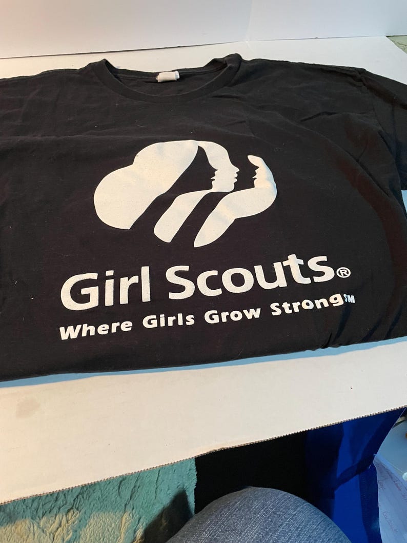 May include: A black short-sleeved t-shirt featuring a white graphic of three stylised female silhouettes. Below the graphic, the text reads Girl Scouts and Where Girls Grow Strong in white lettering. The shirt has a crew neckline and a standard fit.