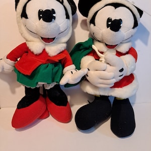 Puede incluir: Un par de juguetes de peluche de Mickey Mouse con tem&aacute;tica navide&ntilde;a. La Mickey Mouse femenina lleva un vestido rojo con una falda verde y ribete de piel blanca. El Mickey Mouse masculino lleva un traje de Pap&aacute; Noel rojo y blanco con una bolsa verde.