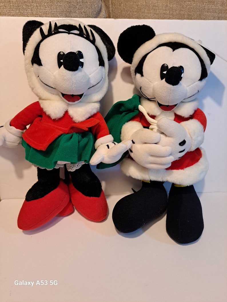 Puede incluir: Un par de juguetes de peluche de Mickey Mouse con tem&aacute;tica navide&ntilde;a. La Mickey Mouse femenina lleva un vestido rojo con una falda verde y ribete de piel blanca. El Mickey Mouse masculino lleva un traje de Pap&aacute; Noel rojo y blanco con una bolsa verde.