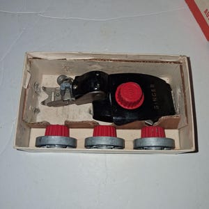 May include: A vintage Singer sewing machine attachment and three red and silver knobs are displayed inside a cardboard box. The attachment is black with a red knob and the Singer logo.