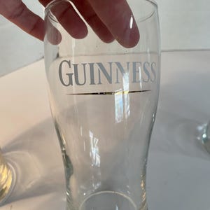 May include: Clear pint glass with the word "GUINNESS" printed in silver. The glass has a curved shape and a thick base. The glass is held by a hand.