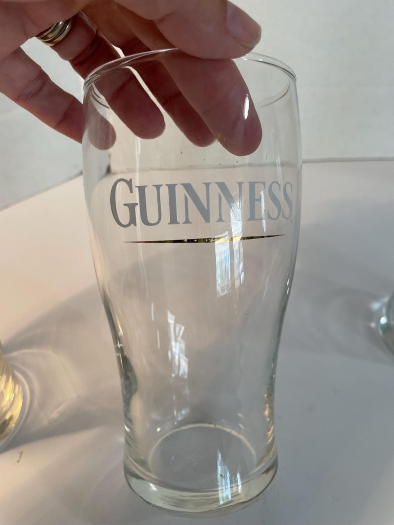 May include: Clear pint glass with the word "GUINNESS" printed in silver. The glass has a curved shape and a thick base. The glass is held by a hand.