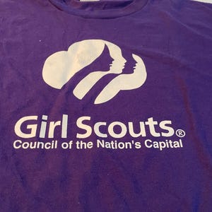May include: A purple t-shirt featuring a white graphic of three stylised silhouettes of faces arranged in a row. Below the graphic, the text reads Girl Scouts in a bold font, followed by the text Council of the Nation's Capital below it.