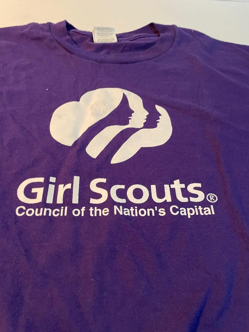 May include: A purple t-shirt featuring a white graphic of three stylised silhouettes of faces arranged in a row. Below the graphic, the text reads Girl Scouts in a bold font, followed by the text Council of the Nation's Capital below it.