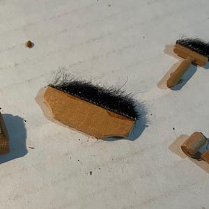 May include: A set of four miniature tools including a small square block with a textured top, a rectangular wooden brush with black bristles, a small T-shaped wooden brush with black bristles, and a tiny wooden roller with a metal handle.