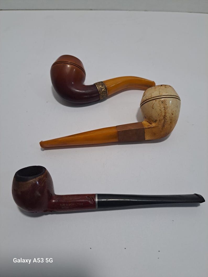 Op de afbeelding: Drie vintage rookpijpen. E&eacute;n pijp is bruin met een gele steel, een andere is wit met een gele steel en de derde is rood met een zwarte steel.