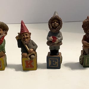 May include: Six small figurines of characters, each standing on a wooden block. The blocks are painted with the letters D, C, K, E, R, E. The characters include figures in hats, hoods, and winter clothing, holding various small items like books and tools.