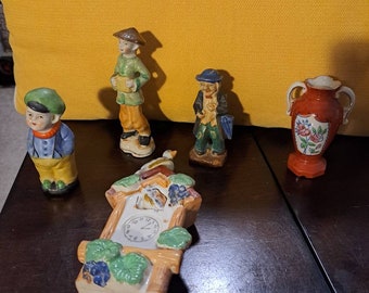 May include: A collection of five vintage porcelain figurines, including a cuckoo clock, a vase with floral designs, and three figures of people. The figures are painted in bright colors and have detailed features.
