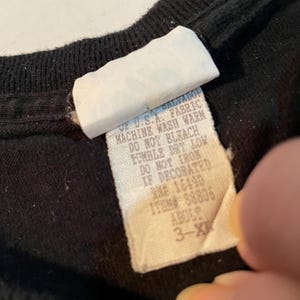 May include: A black garment featuring a white fabric care label with text that reads: OF U.S.A. FABRIC, MACHINE WASH WARM, DO NOT BLEACH, TUMBLE DRY LOW, DO NOT IRON IF DECORATED, RN# 16490, ITEM# 88805, ADULT 3-XL.