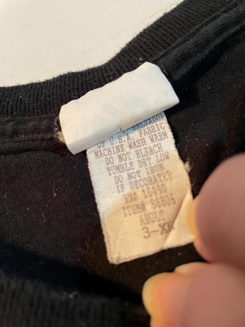 May include: A black garment featuring a white fabric care label with text that reads: OF U.S.A. FABRIC, MACHINE WASH WARM, DO NOT BLEACH, TUMBLE DRY LOW, DO NOT IRON IF DECORATED, RN# 16490, ITEM# 88805, ADULT 3-XL.
