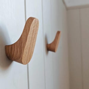 May include: Two wooden wall hooks with a curved design. The hooks are made of light brown wood and are mounted on a white wall. The hooks are designed to hold coats, hats, or other items.
