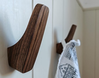 May include: Two wooden wall hooks with a curved design are mounted on a white wall. The hooks are made of dark brown wood with visible grain. One hook has a white and gray patterned towel hanging from it.