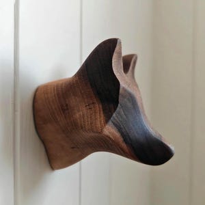 May include: A wooden fox head sculpture fixed to a white wall. The sculpture is crafted from dark and light brown wood, with the grain visible. The fox head is angled, displaying its ears and snout.
