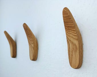 May include: Three wooden wall hooks in a light brown color. The hooks are shaped like a curved blade and are mounted on a white wall.