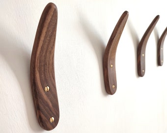 May include: Set of four wooden wall hooks with a curved design. The hooks are made of dark brown wood and are mounted on a white wall. Each hook is secured with two brass screws. The hooks vary slightly in size.