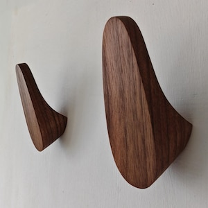 Puede incluir: Dos perchas de pared de madera con un diseño curvo. Los ganchos son de madera marrón oscuro y están montados en una pared blanca. Los ganchos están diseñados para sujetar abrigos, sombreros u otros objetos.