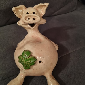 Könnte beinhalten: Eine Keramik-Schweinefigur mit einem grünen Kleeblatt auf dem Bauch. Das Schwein sitzt auf seinen Hinterbeinen, die Vorderbeine sind ausgestreckt. Es hat einen fröhlichen Gesichtsausdruck.