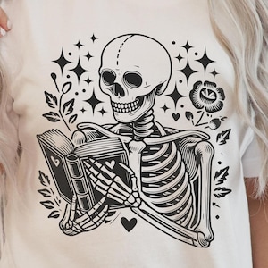 May include: A black and white illustration of a skeleton reading a book with a heart and flowers around it. The skeleton is wearing a shirt with a skull on it.