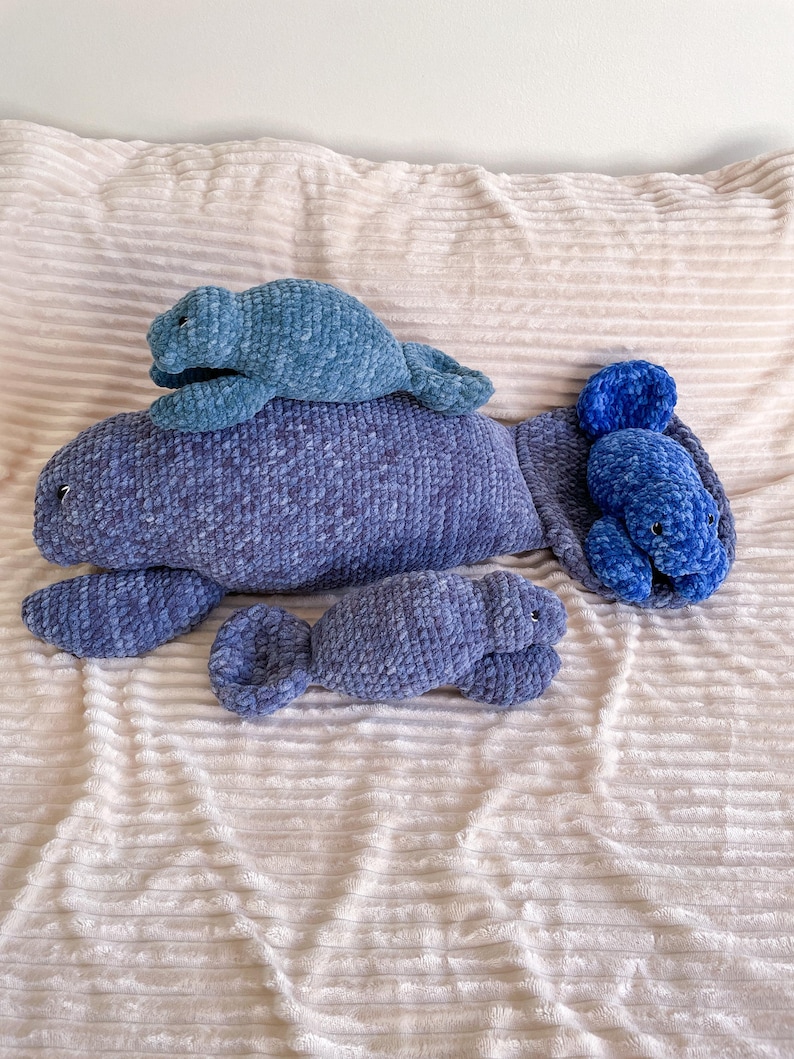 Puede incluir: Tres juguetes de peluche de manat&iacute; de crochet en tonos azules. El manat&iacute; m&aacute;s grande est&aacute; acostado con la cola enroscada. Un manat&iacute; m&aacute;s peque&ntilde;o est&aacute; acostado encima del m&aacute;s grande. El manat&iacute; m&aacute;s peque&ntilde;o est&aacute; acostado de lado con la cola enroscada.