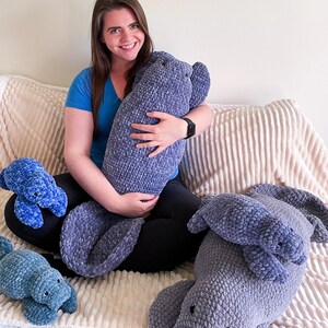 Puede incluir: Una mujer se sienta en un sof&aacute; con cuatro peluches de manat&iacute; tejidos a crochet. El manat&iacute; m&aacute;s grande es gris y los m&aacute;s peque&ntilde;os son azules.