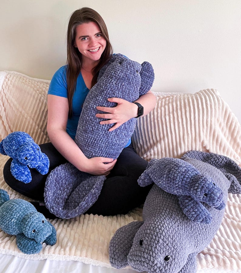 Puede incluir: Una mujer se sienta en un sof&aacute; con cuatro peluches de manat&iacute; tejidos a crochet. El manat&iacute; m&aacute;s grande es gris y los m&aacute;s peque&ntilde;os son azules.