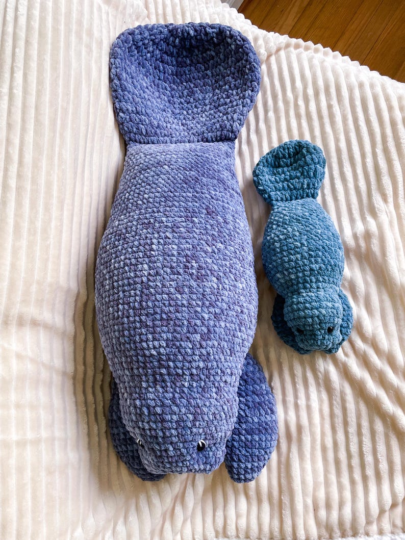 Puede incluir: Dos peluches de manat&iacute; de crochet. El manat&iacute; m&aacute;s grande es azul y el m&aacute;s peque&ntilde;o es azul verdoso. Ambos manat&iacute;es est&aacute;n acostados sobre una manta blanca y esponjosa.