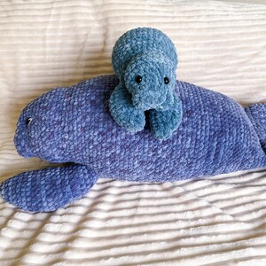 Puede incluir: Un juguete de peluche de manat&iacute; de crochet azul con un juguete de peluche de tortuga de crochet azul m&aacute;s peque&ntilde;o encima. Ambos juguetes est&aacute;n sentados en una manta blanca y esponjosa.